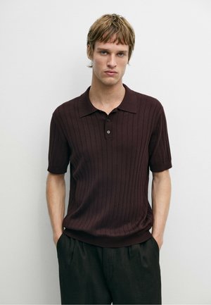 Junger Mann mit hellbraunen Haaren, der ein kurzärmeliges dunkelbraunes, geripptes Poloshirt und schwarze Hose trägt, steht vor einem schlichten weißen Hintergrund.