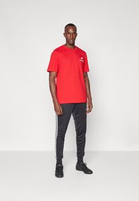Rotes T-Shirt mit kurzen Ärmeln, aus Baumwollmaterial, mit einem Logo auf der Brust. Kombiniert mit schwarzen Jogginghosen, die an den Seiten drei weiße Streifen haben.