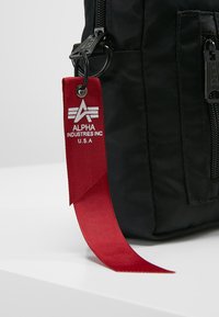 Musta nailonilaukku vetoketjulla, jossa on näkyvä punainen kangastunnus, jossa lukee "ALPHA INDUSTRIES INC." ja siivillä varustettu logo.