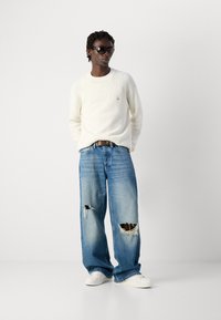 Man in een wit gebreid sweater, met een wijd uitlopende ripped spijkerbroek, witte sneakers en donkere zonnebril, staande tegen een effen lichte achtergrond.