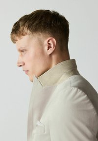 Jeune homme aux cheveux courts et bruns porte une veste beige à col montant, tourné vers la gauche sur un fond clair uni.