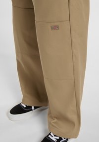 Beiga arbetsbyxor med lös passform, med en logo i lappstil på låret, tillverkade av slitstarkt tyg, och ihop med svarta sneakers.