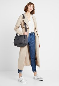 Gray fabric shoulder bag with multiple compartments and a detachable strap, paired with a beige trench coat and denim jeans. White sneakers.