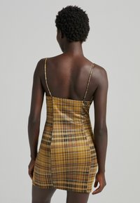 Abito fitted a quadri gialli e marroni con spalline sottili, zip sul retro e texture del tessuto liscia. Lunghezza sopra il ginocchio.