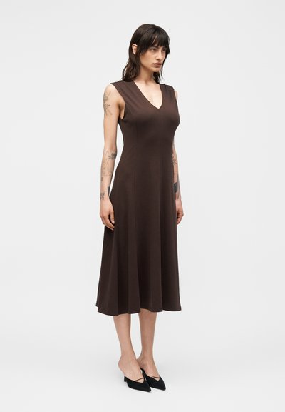 Γυναίκα με σκούρα μαλλιά, ντυμένη με αμάνικο καφέ μίντι φόρεμα και μαύρες μυτερές γόβες, στέκεται μπροστά σε απλό ανοιχτό φόντο.