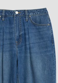 Close-up of blue denim jeans waist with button, zipper, belt loops, front pocket, and contrast stitching on a light background.