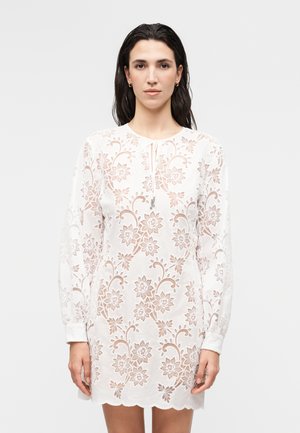 Žena s dlhými tmavými vlasmi má na sebe biele kvetinové čipkované šaty s dlhými rukávmi a zubatým okrajom, stojí pred jednoduchým svetlým pozadím.