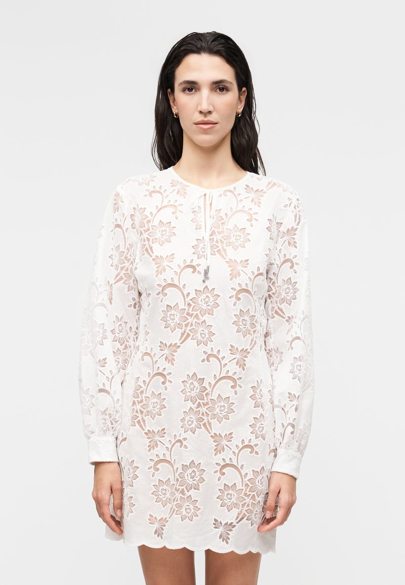 Γυναίκα με μακριά σκούρα μαλλιά φοράει λευκό φόρεμα με φλοράλ δαντέλα, μακριά μανίκια και οδοντωτό τελείωμα, στέκεται μπροστά από απλό ανοιχτό φόντο.