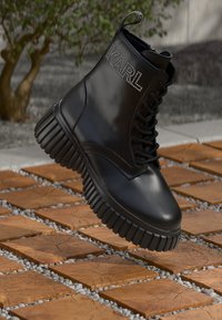 Črne usnjene vojaške čevlje z debelejšim režastim podplatom in "KARL" iz rhinestones na strani, lebdeče nad lesenim blokom na pločniku na prostem.