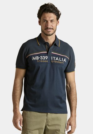 Hombre con cabello corto y oscuro, vistiendo una camiseta polo azul marino con el texto "MB-339 ITALIA frecce tricolori", combinada con pantalones cargo beige.