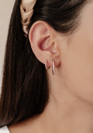 Roségoldene Creolen mit einem schlichten Design, die an einer Seite kleine, klare Kristalle aufweisen. Nahaufnahme der Ohrringe, die im Ohr getragen werden.