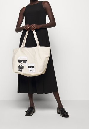 Beige Canvas-Umhängetasche mit grafischen Drucken von zwei Figuren, die Sonnenbrillen tragen. Sie hat lange Griffe für einen bequemen Transport.