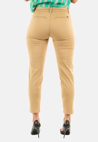 Pantalones beige ajustados con corte slim, con bolsillos traseros y una textura suave. Combinados con tacones altos negros. Parte superior de rayas de colores brillantes visible.