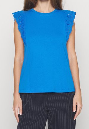 Femme portant un haut sans manches bleu vif avec des volants en dentelle ajourée sur les épaules et un pantalon noir à fines rayures.