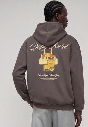 Homme aux cheveux tressés portant un sweat à capuche marron foncé avec "Deeply Rooted" et un graphisme de chariot à hot-dogs de Brooklyn dans le dos.