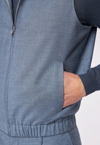 Hellblauer Jumpsuit aus strukturiertem Stoff mit Reißverschluss, zwei Seitentaschen und elastischem Bund. Verfügt über kontrastierende dunkelblaue Ärmelakzente.