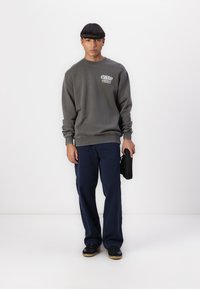 Gray sweatshirt featuring a brand logo on the chest, paired with navy blue pants and black sneakers. The fabric appears soft and relaxed.