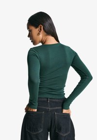 Langärmliges, enganliegendes Oberteil aus dunklem grünen Baumwollstoff, mit rundem Ausschnitt und glatter Textur, kombiniert mit dunklen Jeans.
