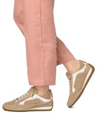 Baskets en suède beige avec des accents blancs, présentant un design perforé, une semelle en caoutchouc et un laçage à l'avant, associées à un pantalon en lin rose clair.