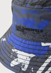 Szürke alapon kék és fehér absztrakt foltmintás halászkalap. Texturált anyag, az oldalán hímzett logóval.