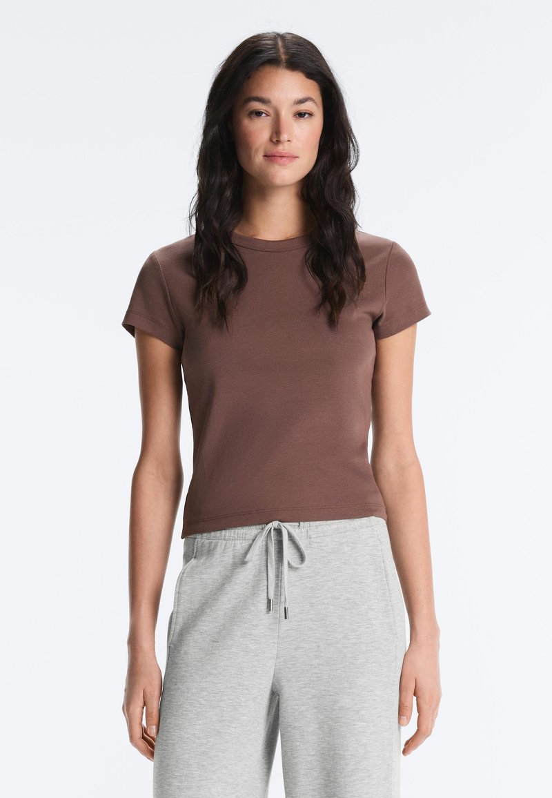 Femme aux longs cheveux foncés, portant un t-shirt ajusté marron à manches courtes et un pantalon gris clair à cordon, debout sur un fond blanc.