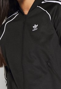 Svart idrottszip-jacka i mjukt tyg, med vita ränder på axlarna, sidofickor och en broderad logotyp på bröstet.