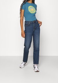 Camiseta azul de manga corta con un gráfico de girasol circular, combinada con jeans oscuros de tiro alto y zapatillas blancas.