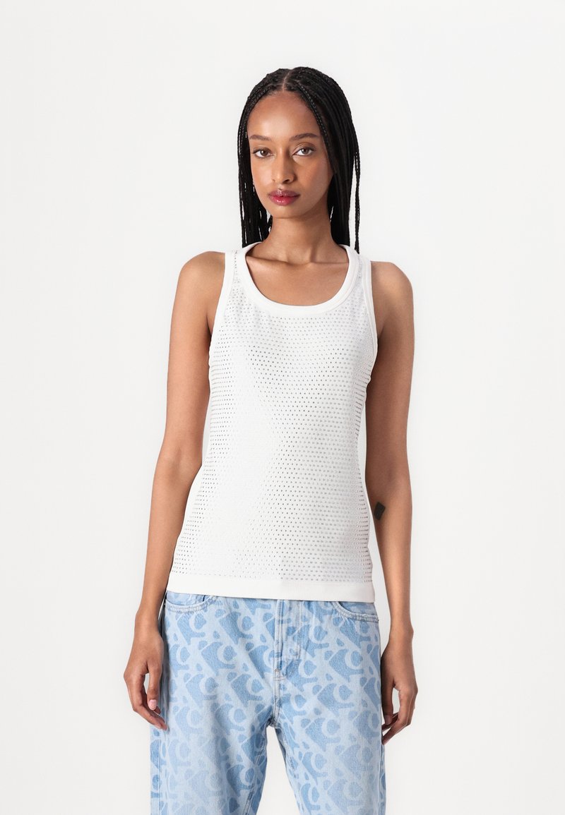 Weißes ärmelloses Tanktop mit Netzstruktur und festem Saum. Getragen mit hellblauen, gemusterten Jeans, die ein dezentes Logo-Design zeigen.