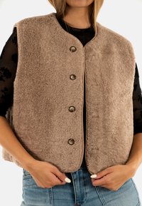 Gilet in finta pelliccia shearling marrone con scollatura rotonda, cinque bottoni sul davanti e tasche, indossato sopra una maglia nera a maniche lunghe.