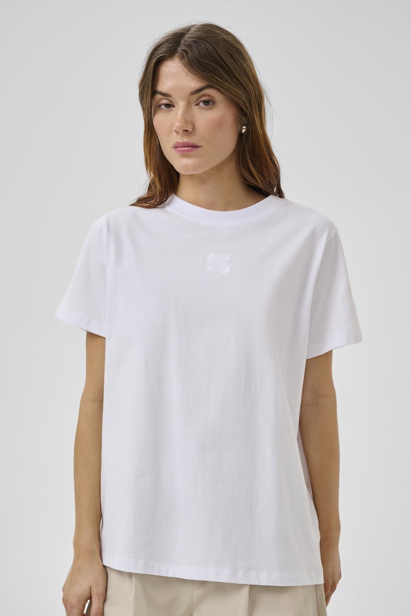 Femeie care poartă un tricou alb simplu cu mâneci scurte și pantaloni bej, stând în fața unui fundal gri neutru.
