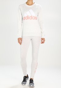 Sweatshirt gris clair avec logo Adidas blanc et rose, associé à des leggings gris clair assortis. Chaussures de sport noires avec accents réfléchissants.