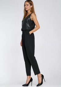 Czarny cekinowy krótki top w połączeniu z czarnymi spodniami z wysokim stanem. Modelka w czarnych szpilkach, ręce w kieszeniach, neutralne tło.