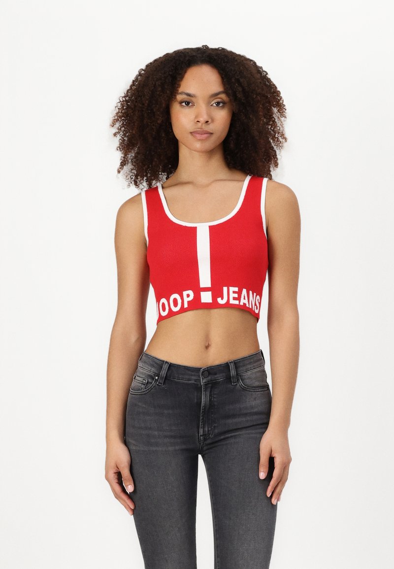 Rød crop-top med scoop-hals og hvit tekst som sier "DOOP JEANS", kombinert med mørke skinny jeans. Stoffet har en ribbestrikket tekstur.