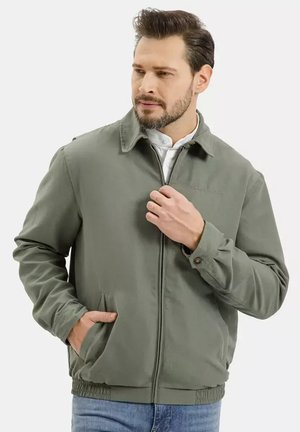 Mężczyzna z brodą w zielonej kurtce zapinanej na zamek i jasnoniebieskich dżinsach, ręka w kieszeni, patrzący na bok na tle białego tła.