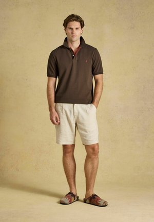 Giovane uomo in piedi che indossa una polo marrone scuro, pantaloncini beige e scarpe slip-on con motivo su uno sfondo beige uniforme.