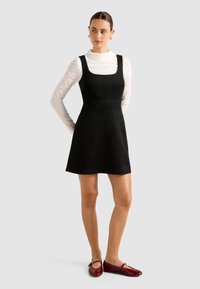 Femme portant une robe noire sans manches en ligne A, un haut blanc texturé à manches longues, des boucles d'oreilles en or et des ballerines rouges Mary Jane, debout.