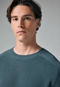 Pull vert côtelé avec un col rond et un tissu texturé, présentant des lignes horizontales subtiles et une coupe ajustée, idéal pour un look décontracté.