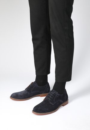 Marineblauwe suède schoenen met een gladde textuur, voorzien van een ronde neus, veters en een rubberen zool, gecombineerd met zwarte broeken en sokken.