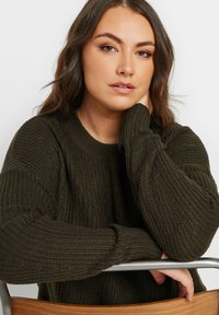 Mörkgrön stickad tröja med ribbad textur och rund halsringning. Har långa ärmar och en avslappnad passform, använd när man vilar på en stol.