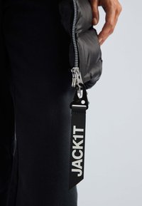 Zwarte pufferjas van glanzende stof, met een metallic rits en een stoffen label met de tekst "JACK1T". De textuur lijkt glad en geïsoleerd.