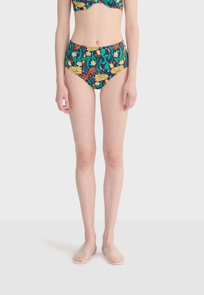 Femme portant un bas de bikini taille haute à fleurs et un haut assorti aux fleurs multicolores, debout pieds nus sur un fond blanc.