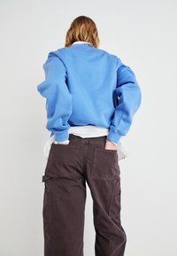Lyseblå hettegenser med senkede skuldre, kombinert med mørkebrown cargo-bukser med store lommer, sett bakfra.