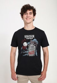 Camiseta negra de algodón con un diseño gráfico colorido que incluye una tumba, murciélagos y una casa, etiquetada con "STRANGER THINGS" y "HELLFIRE FOREVER."