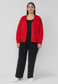 Rode gebreide vest met lange mouwen, V-hals, knoopsluiting aan de voorkant en twee zakken aan de voorkant, gecombineerd met een zwarte broek en witte sneakers.