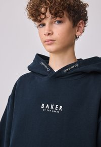 Marineblå hættetrøje lavet af blødt stof med et frontlogo "BAKER BY TED BAKER" i hvid. Inkluderer en snøre i hætten med brandede detaljer.