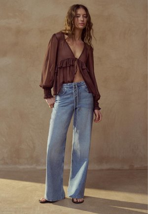 Femeie purtând o bluză maro transparentă cu volane și blugi largi, de talie înaltă, de culoare albastru deschis, stând în picioare în fața unui perete bej simplu.