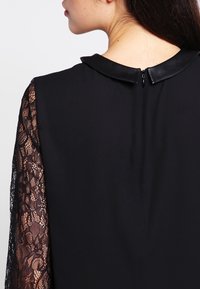 Abito nero con manica lunga in pizzo su un lato, corpo nero uniforme e colletto in finta pelle con chiusura a zip sul retro.
