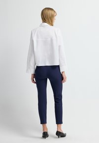 Camisa blanca corta de botón con mangas largas, combinada con pantalones ajustados azul marino y tacones negros, vista desde atrás. Textura de tela suave.