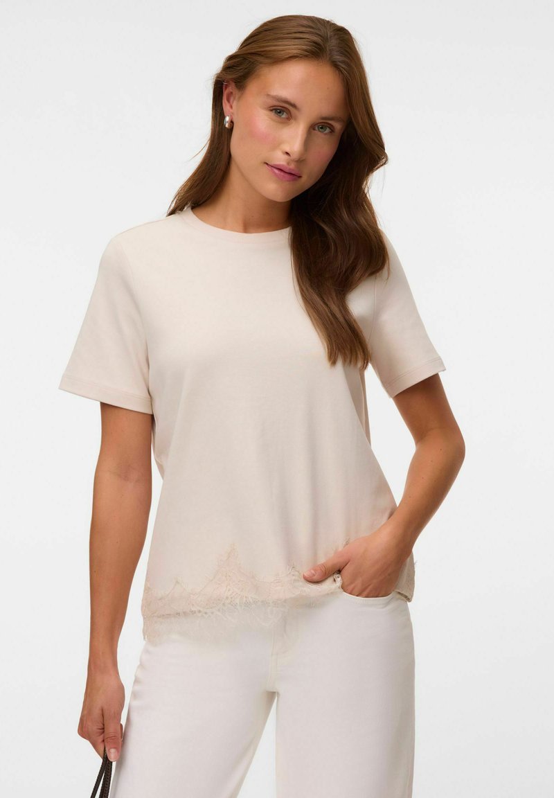 Femme aux longs cheveux bruns portant un haut beige clair à manches courtes avec un ourlet en dentelle et un pantalon blanc, tenant un sac à main noir à bandoulière.