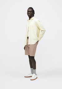Lichtgele blouse met lange mouwen en knopen, lichtbruine shorts, witte sneakers, grijze sokken. Model staat met handen in de zakken. Effen achtergrond.
